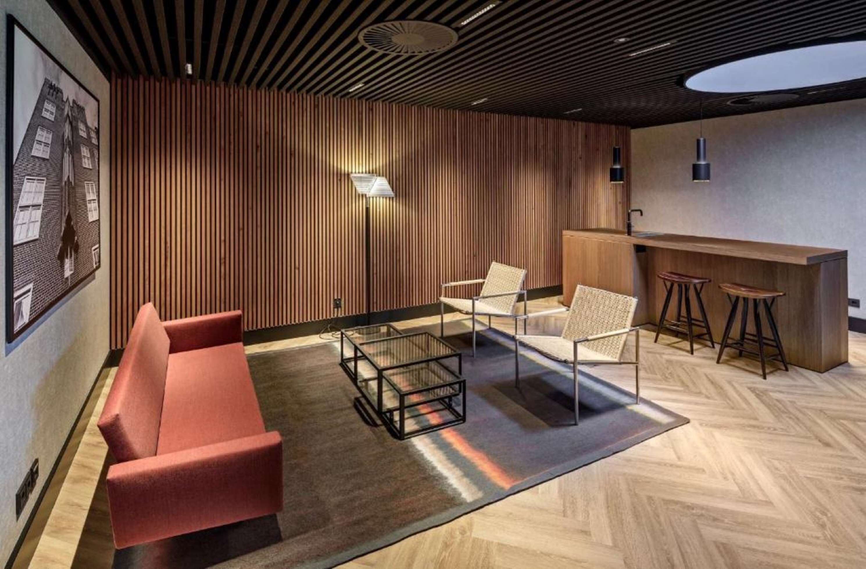 A modern interior with a wooden panel wall, a red sofa, a glass coffee table, and a bar area with stools.