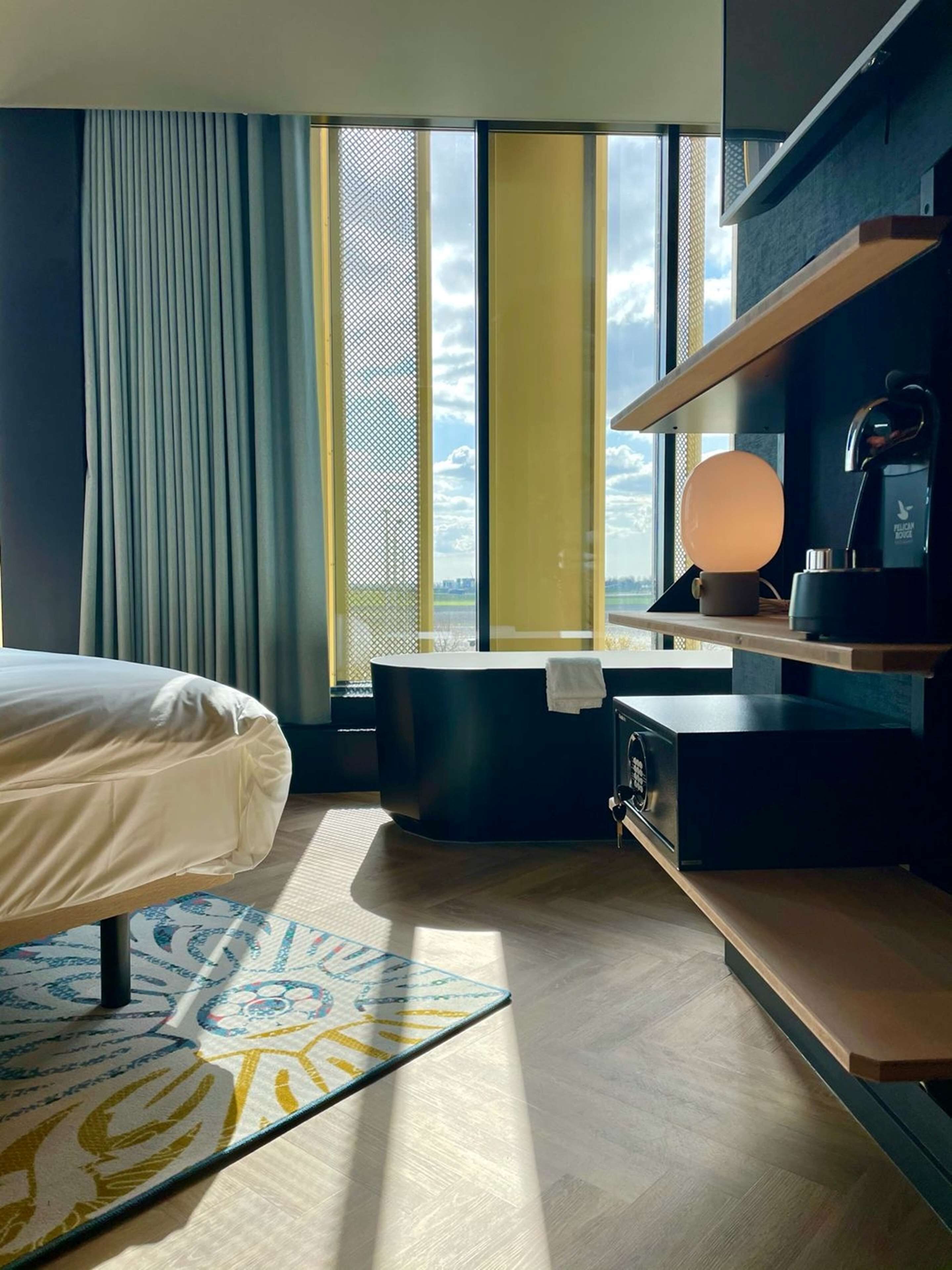 A hotel room with a bed, a desk, a chair, a small table, a lamp, and a view of the outdoors through large windows.