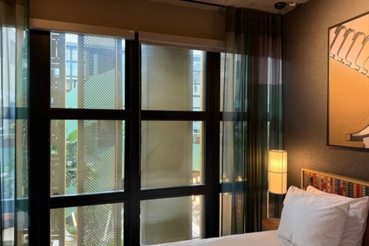 A hotel room with a large window, a bed with white linens, a nightstand with a lamp, and a wall-mounted artwork.