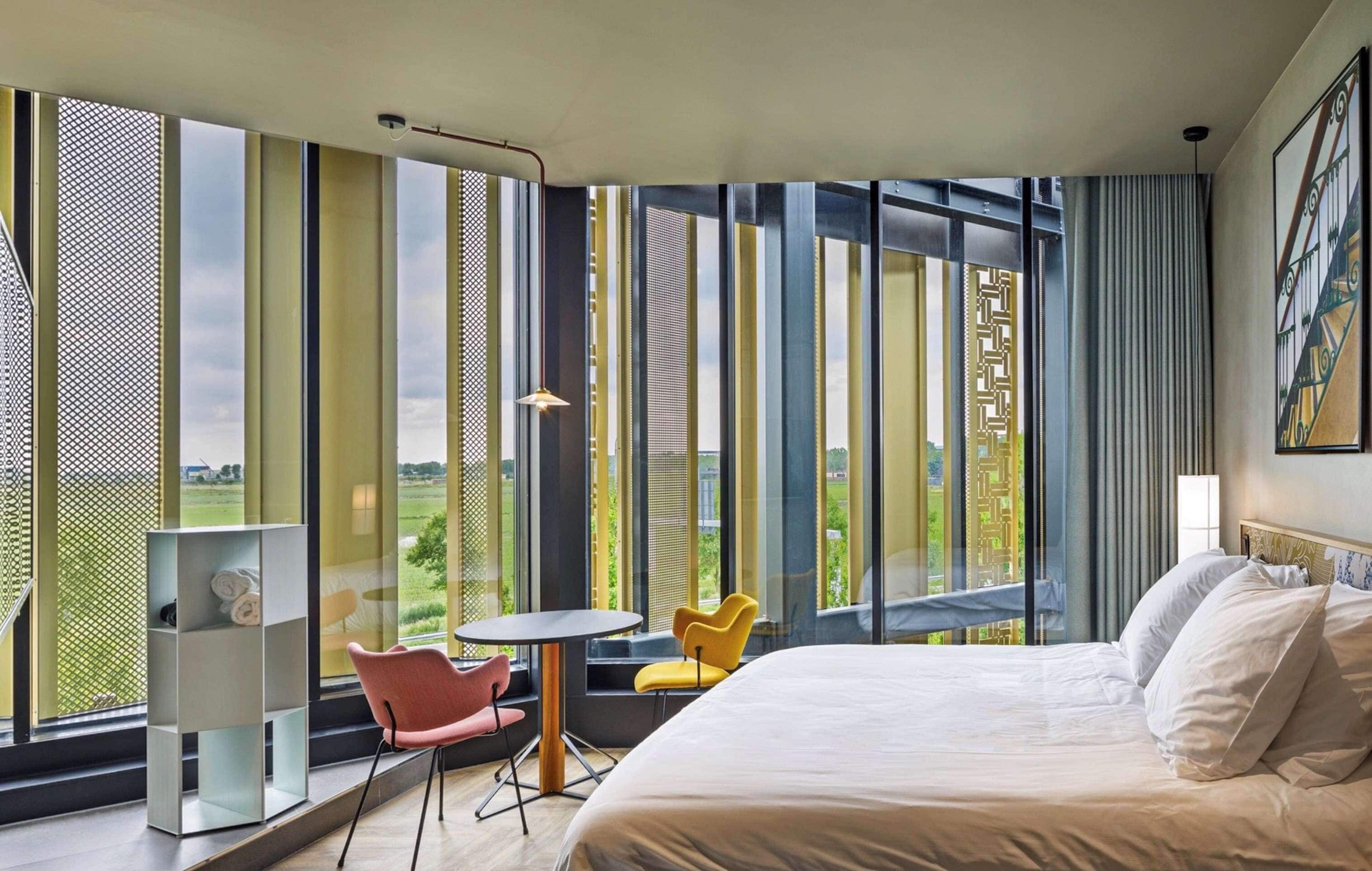 A modern hotel room with a large bed, a small table, two chairs, and a view of the outdoors through large windows.