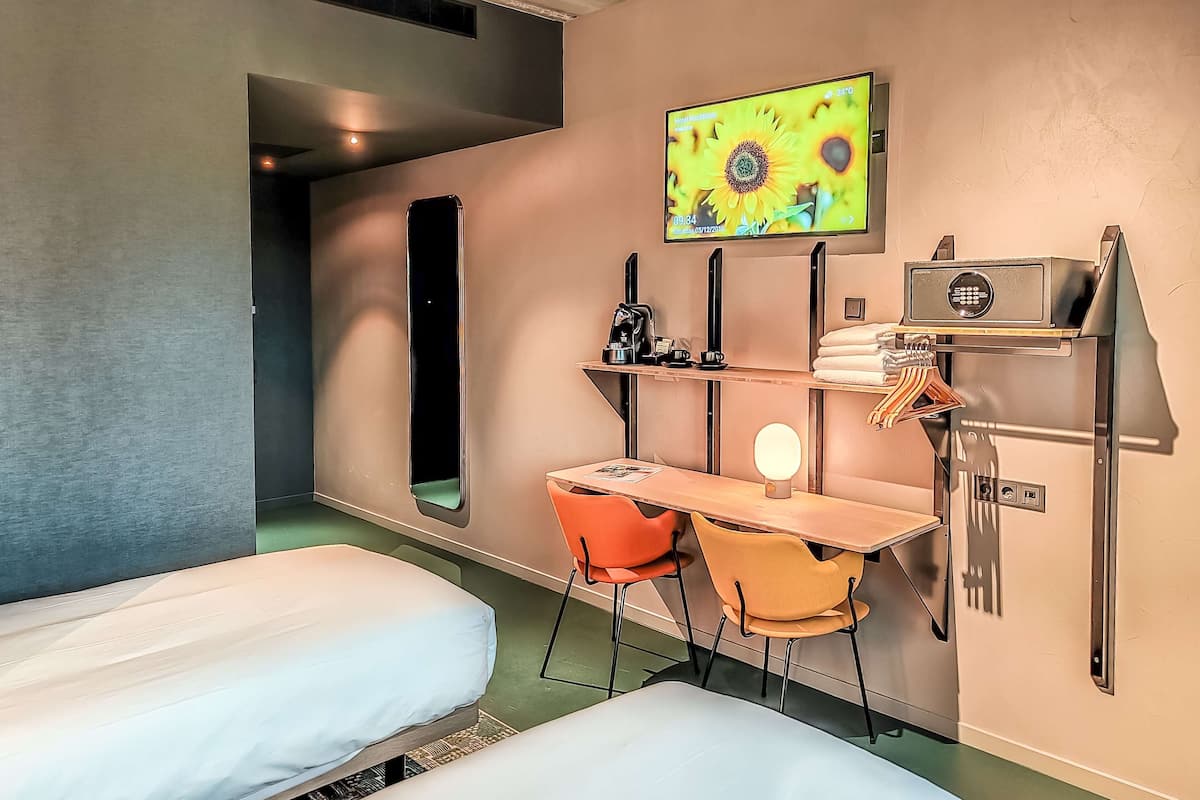 A hotel room with two beds, a desk with a chair, a TV on the wall, a coffee machine, and a safe.