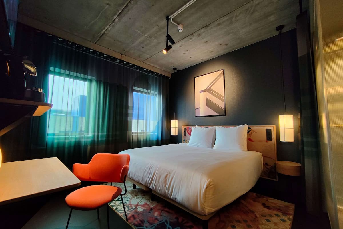 A modern hotel room with a large bed, orange chairs, a desk, and a view of the city through large windows.
