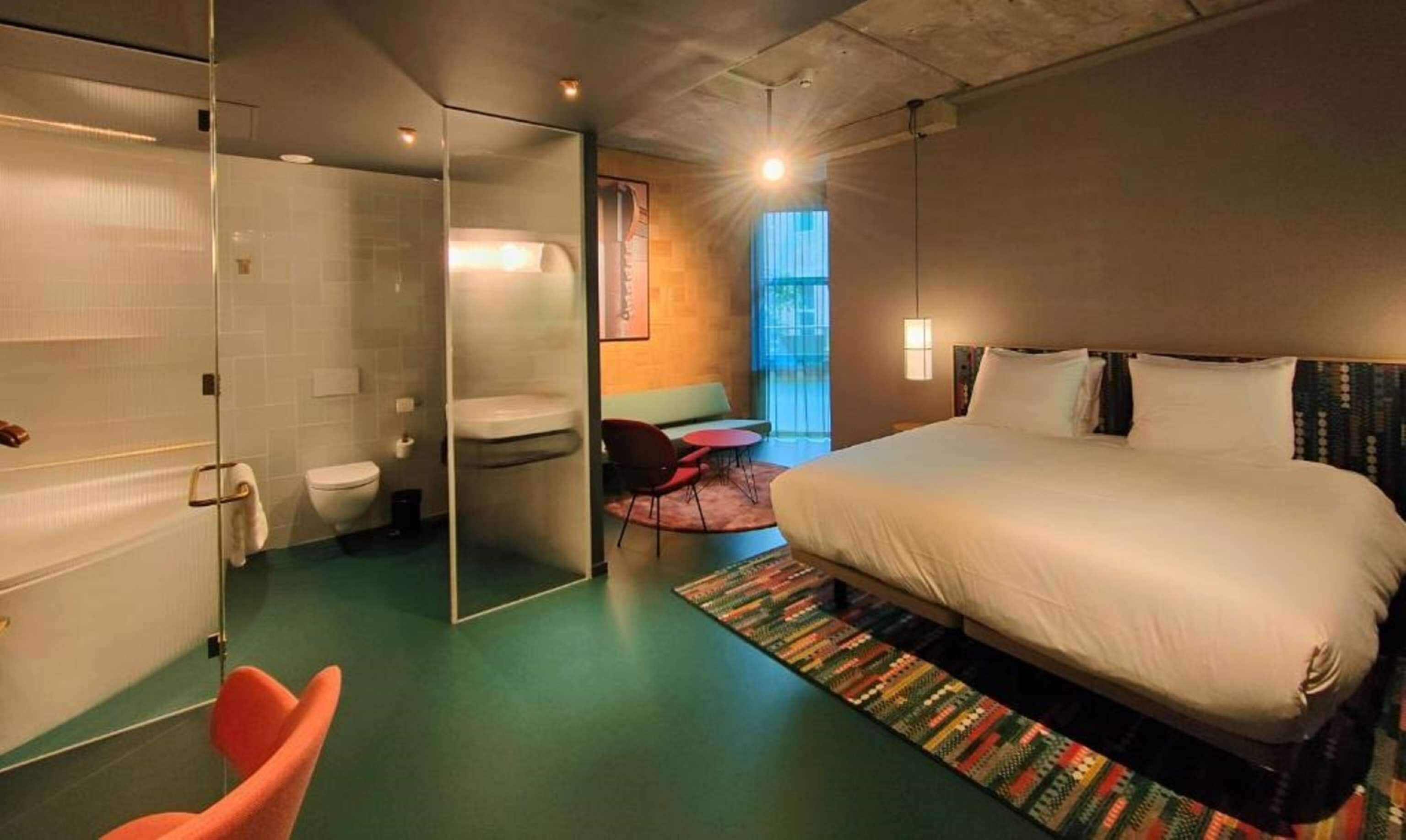 Ein modernes Hotelzimmer mit einem großen Bett, einer Duschkabine aus Glas, einer kleinen Sitzecke und Blick ins Freie.