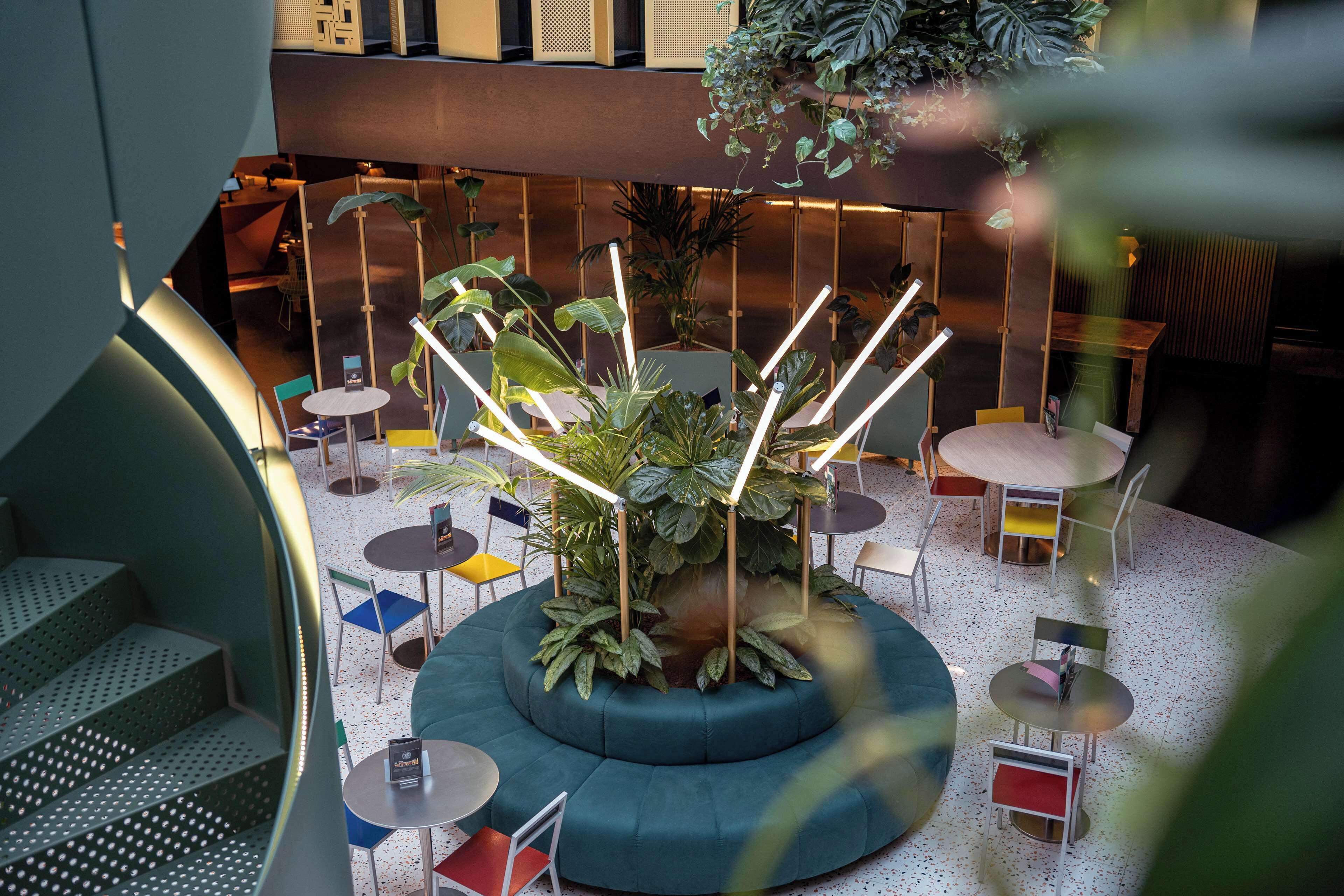 A modern indoor space with a central seating area surrounded by tables and chairs, a staircase, and a variety of plants.