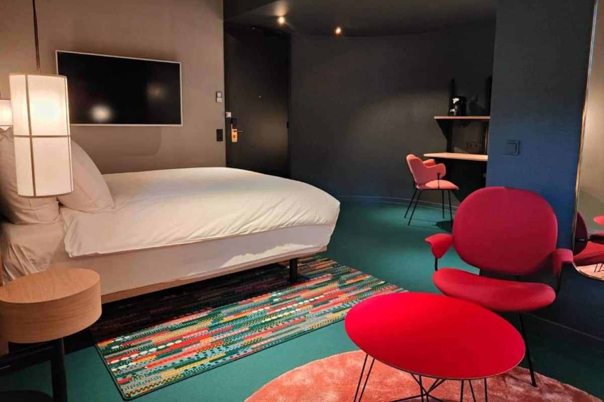 A modern hotel room with a large bed, a desk, and red chairs.