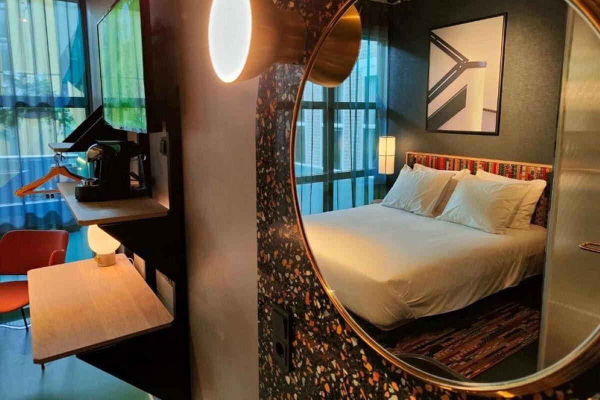 A hotel room with a bed, a desk with a computer, a coffee machine, and a large mirror.