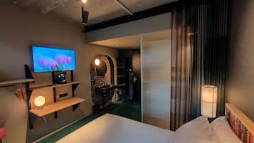 A hotel room with a bed, a TV on the wall, a desk with a chair, and a bathroom visible through a glass partition.