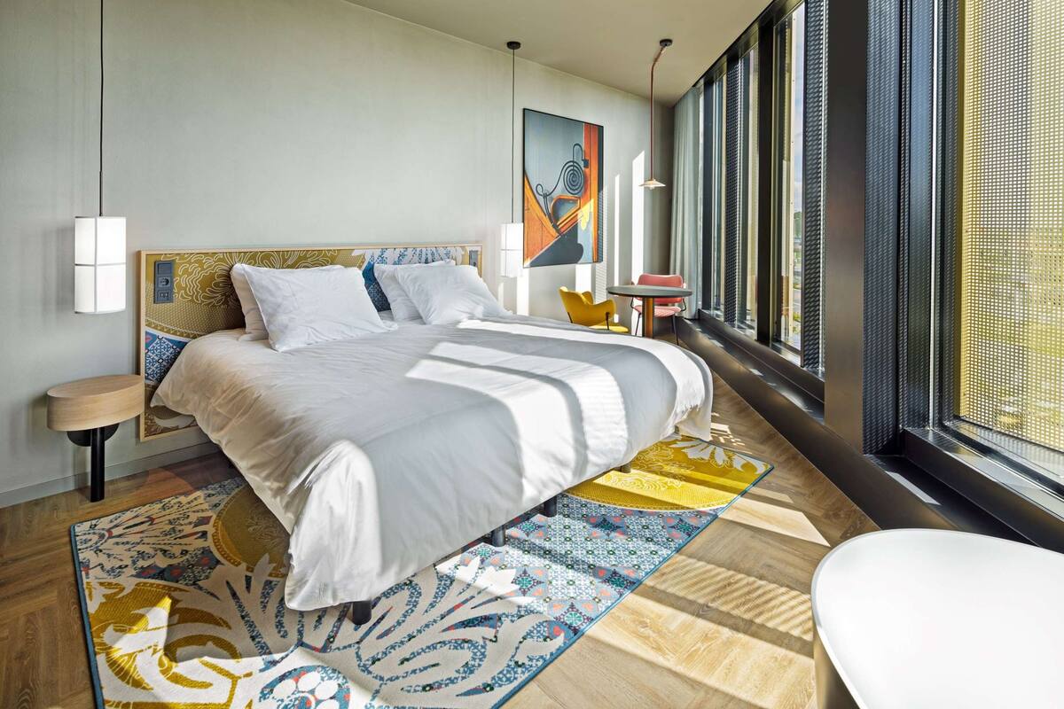 A modern hotel room with a large bed, a small table, a chair, and a large window with a view.