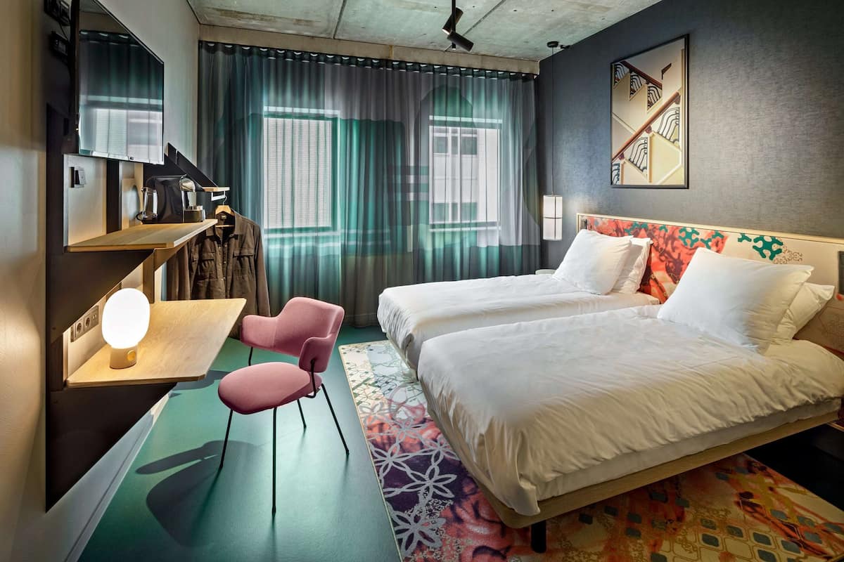 A hotel room with two beds, a desk with a lamp, a chair, and a large window with curtains.