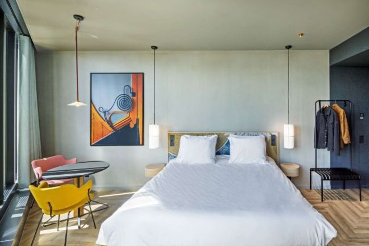A modern hotel room with a large bed, a desk, a chair, and a painting on the wall.