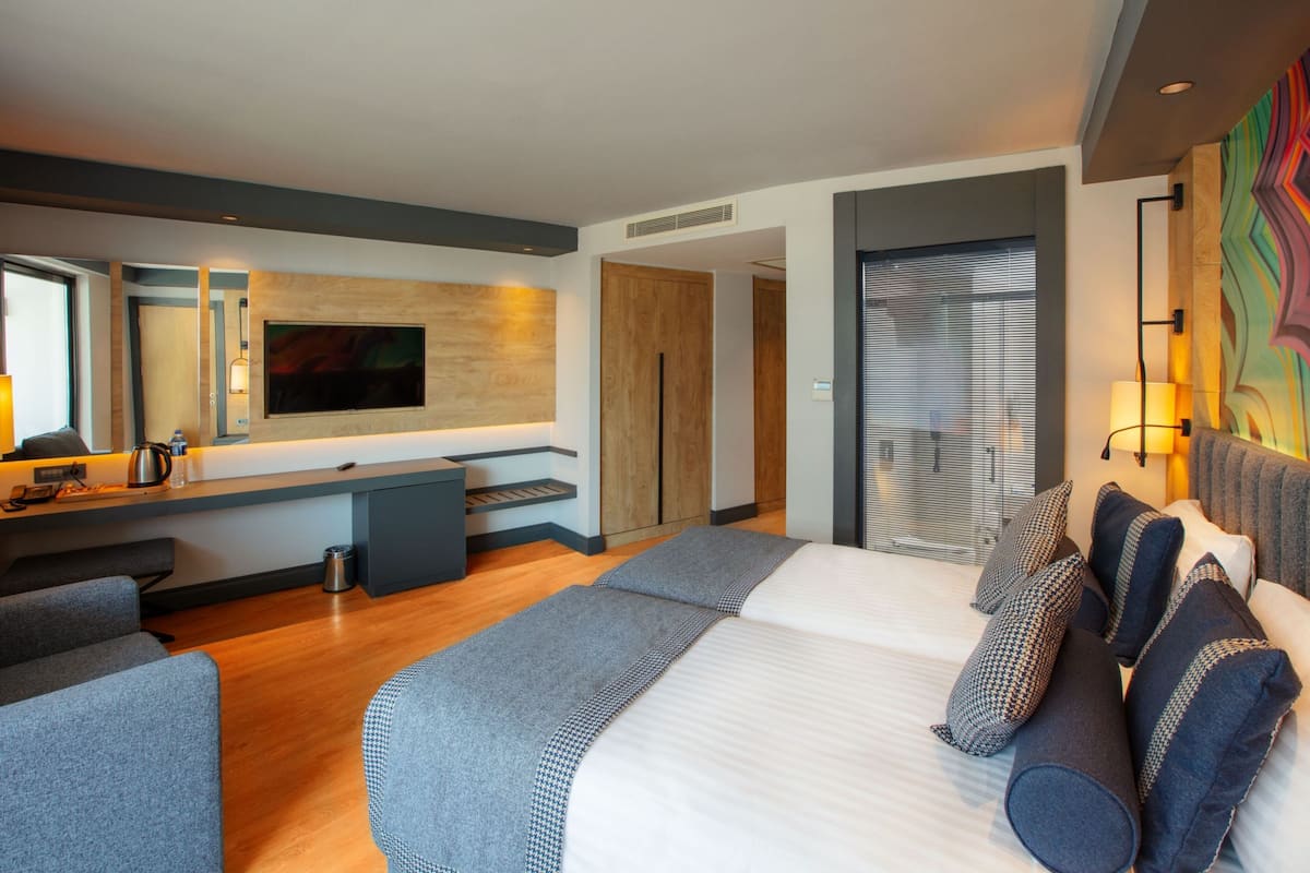 A modern hotel room with a large bed, a desk with a flat-screen TV, a sofa, and a large window with blinds.
