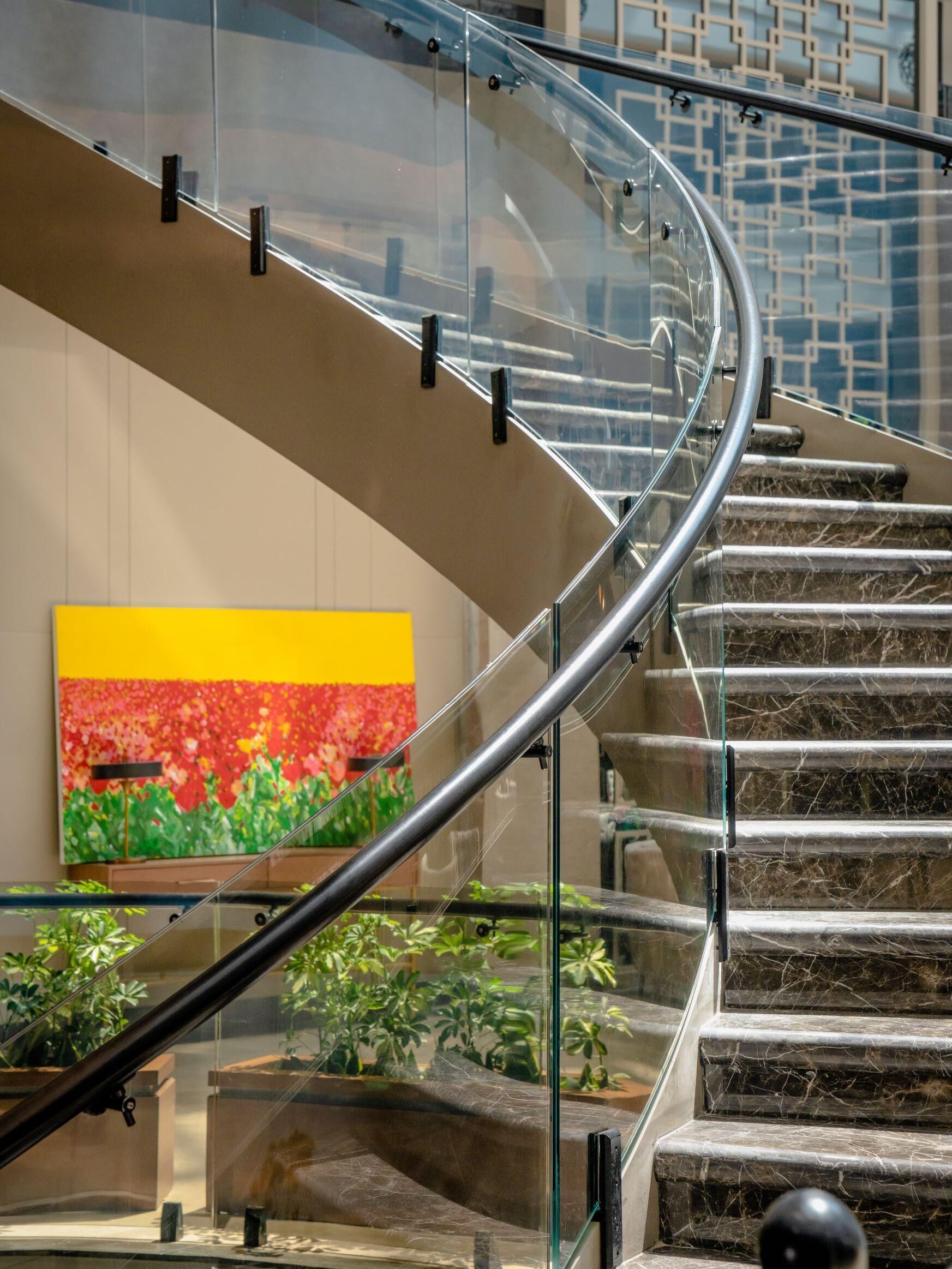A curved glass staircase with metal railings, a yellow and red painting on the wall, and potted plants at the bottom.