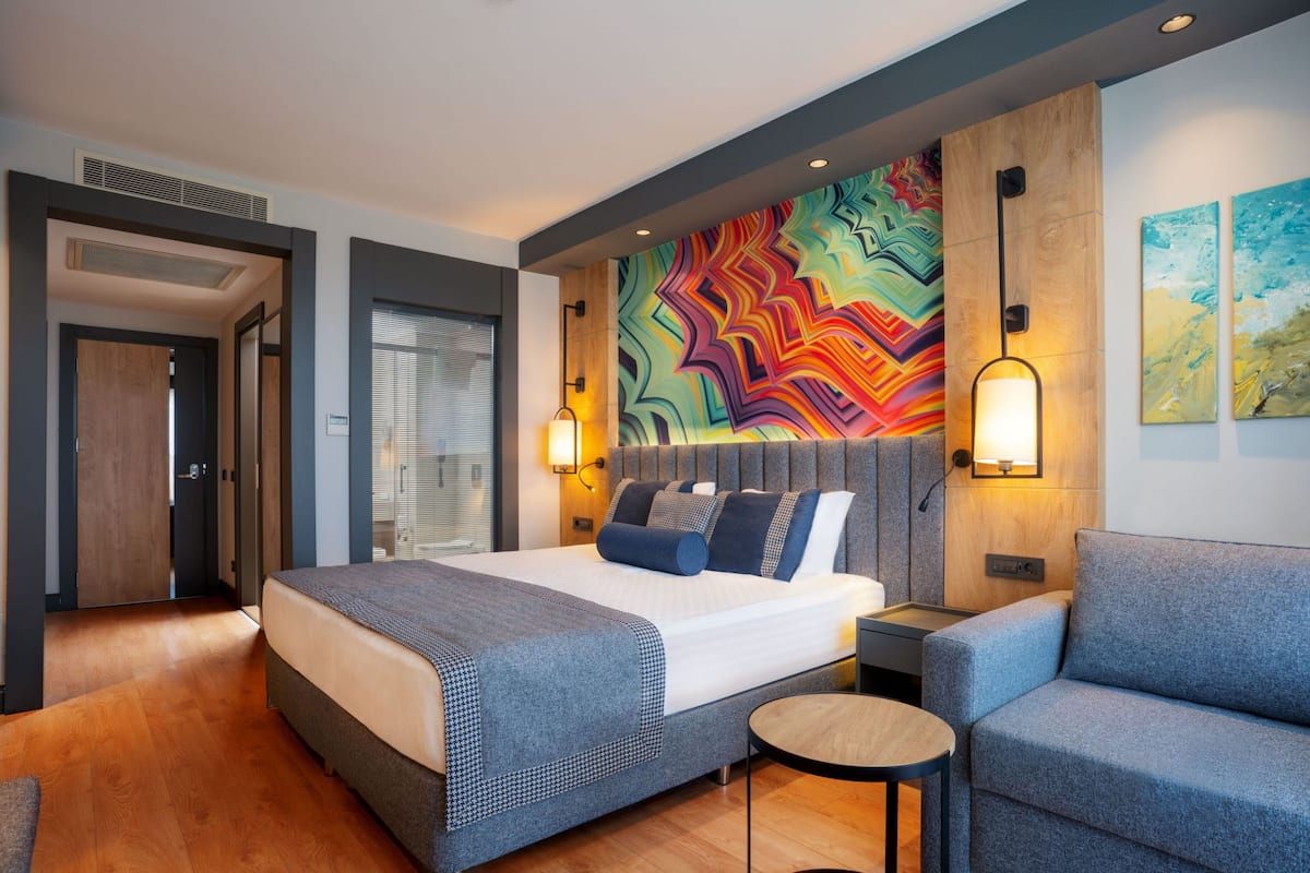 A modern hotel room with a large bed, a small round table, and a colorful abstract wall art.