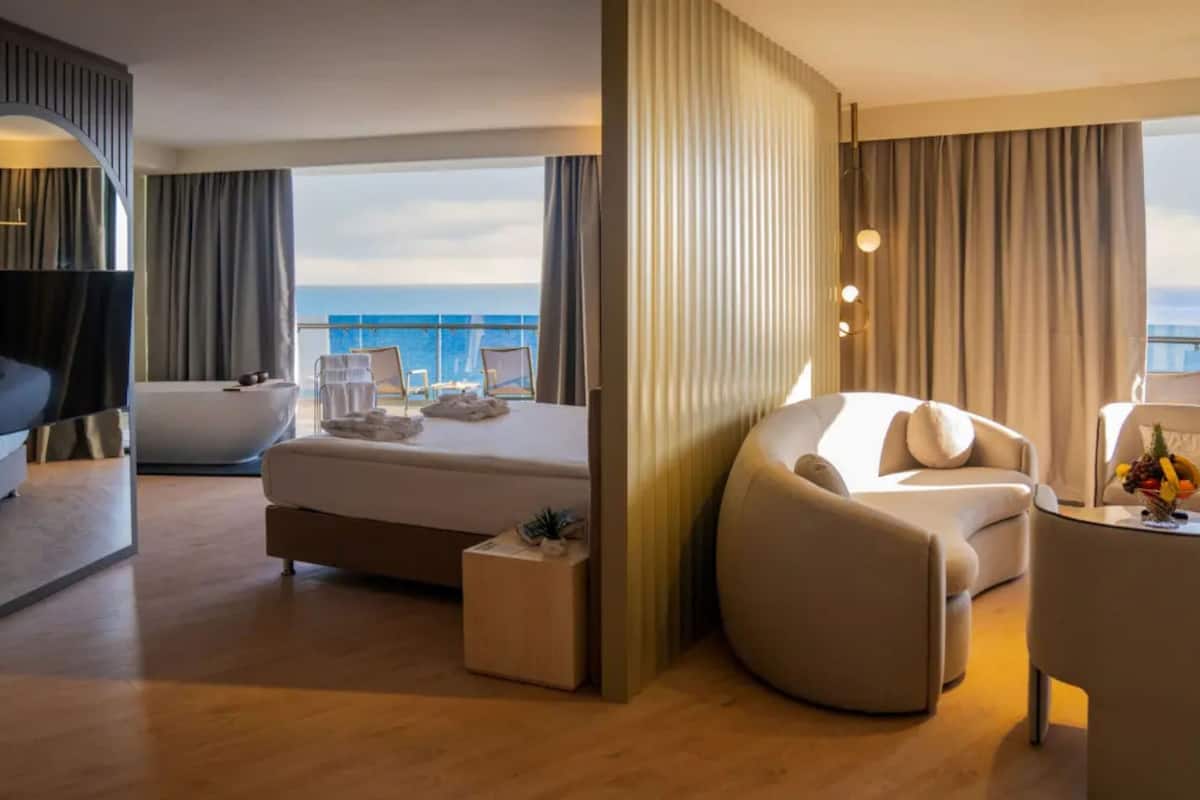 A modern hotel room with a large bed, a sofa, a small table, and a view of the ocean.