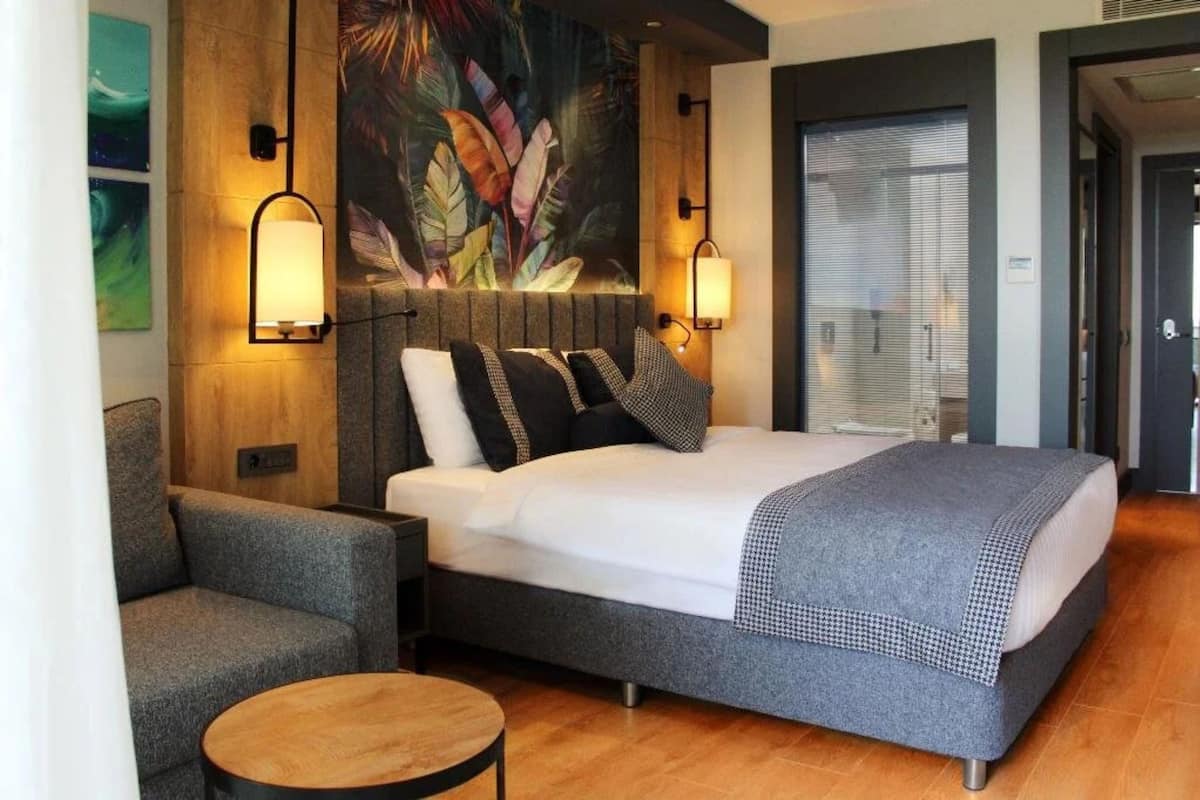 A hotel room with a large bed, a grey sofa, a round wooden table, and a large wall mural.