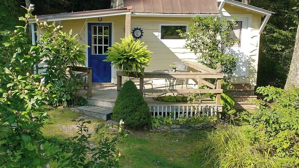 Una casita de un piso con puerta azul, rodeada de exuberante vegetación y cerca blanca de piquete.