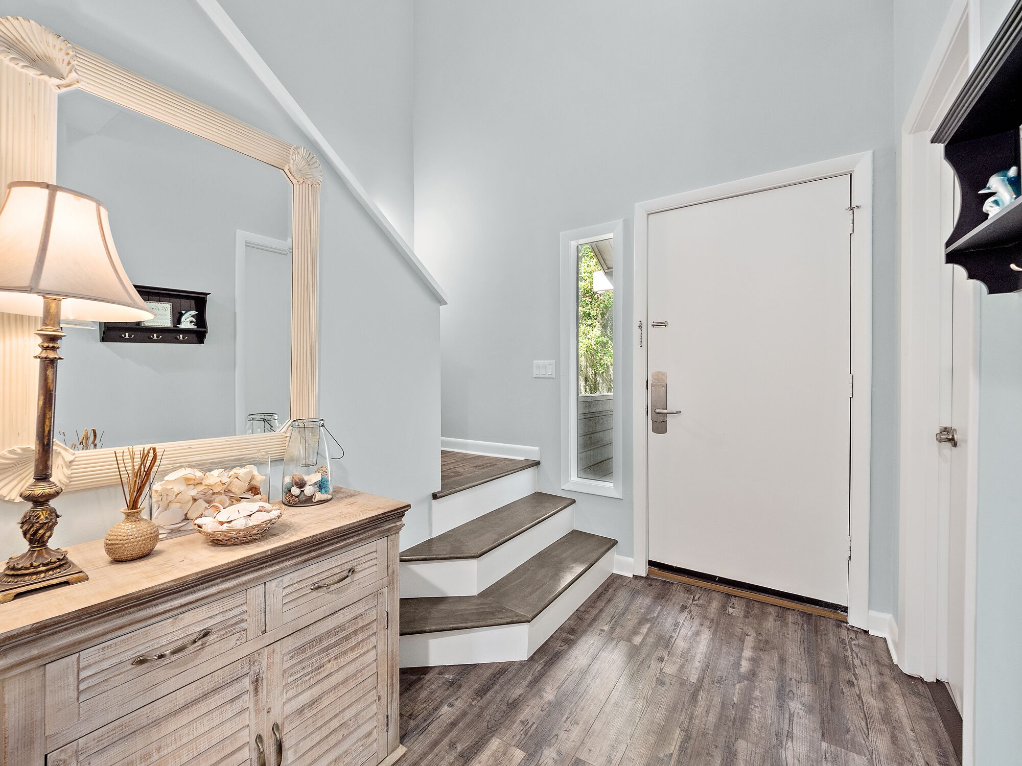 A hallway with a wooden dresser, a mirror, and a lamp. There are stairs leading to a door with a window.