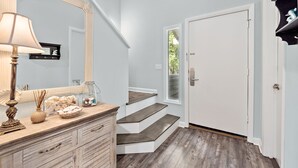 Een gang met een houten dressoir, een spiegel en een lamp. Er zijn trappen die naar een deur met een raam leiden.