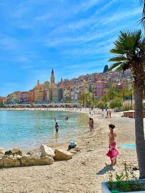 A beach with people swimming and walking, a palm tree, and colorful buildings in the background. - 3 rooms by the water, 30 m from the park beaches (Roquebrune-Cap-Martin)