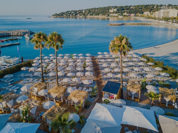 A beachside resort with numerous sun loungers and umbrellas, a wooden deck, and a view of the sea and distant buildings.