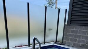 A small outdoor hot tub with a metal safety rail, surrounded by a tiled deck and a glass wall.