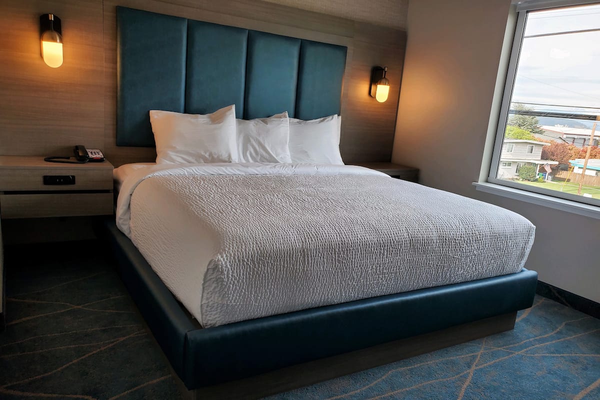 A hotel room with a large bed, a headboard, bedside lamps, a nightstand with a phone, and a window with a view of the outside.