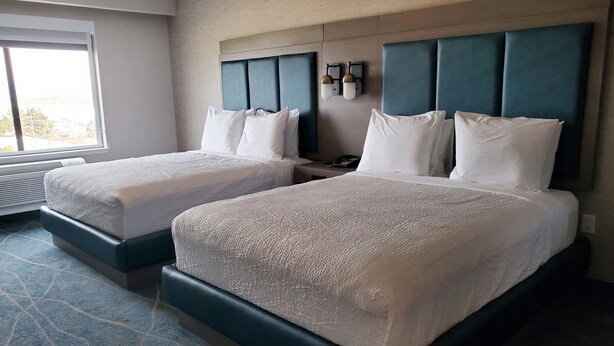 A hotel room with two beds, a large window, and a wall-mounted light fixture.