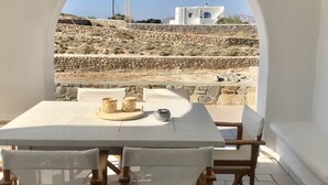 Une table entourée de chaises, avec vue sur un mur de pierre et des bâtiments, le tout visible à travers une fenêtre en arc de cercle.