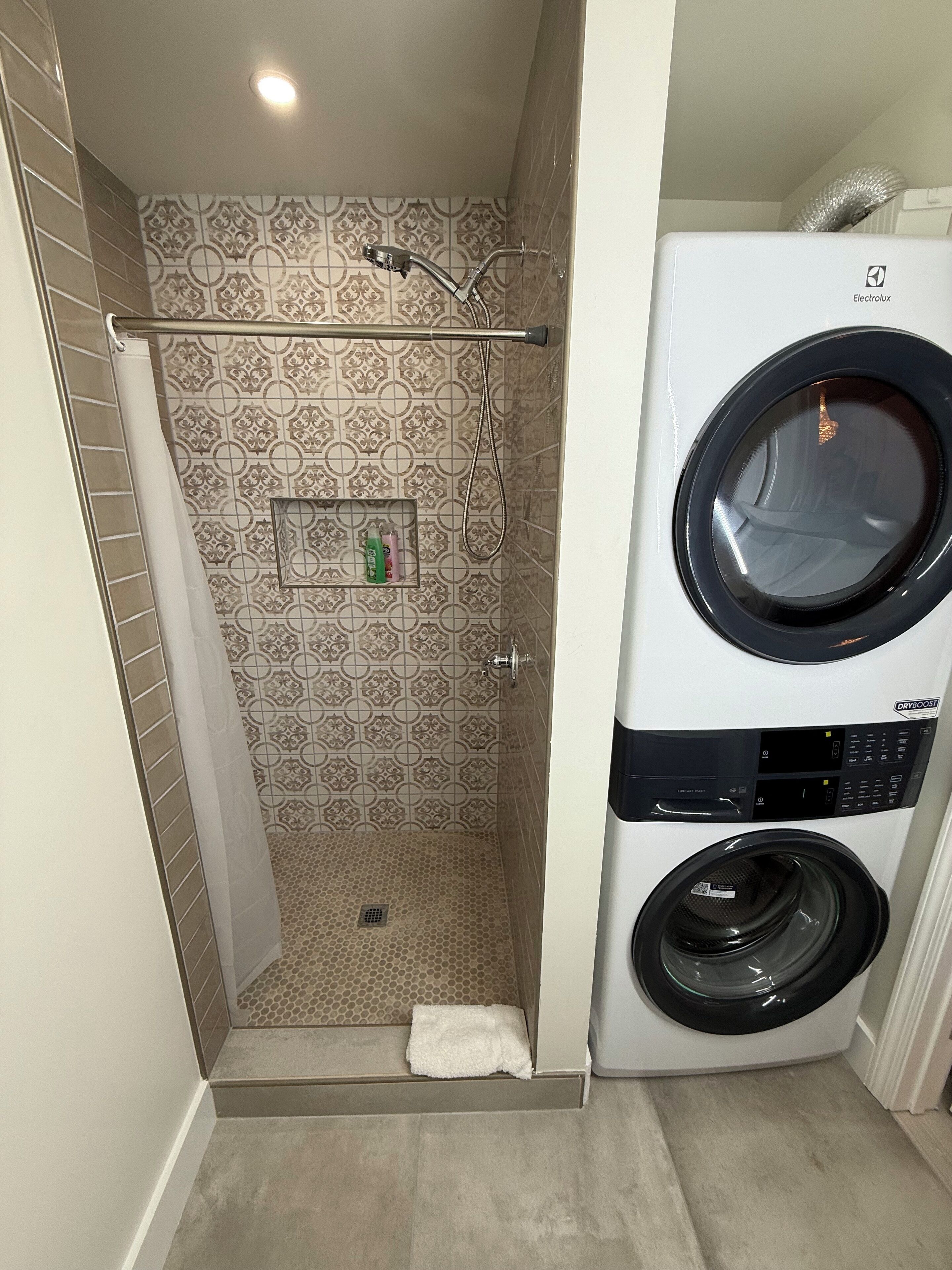 Un lavadero pequeño con lavadora y secadora, una zona de ducha con azulejos estampados y un estante con artículos de aseo.