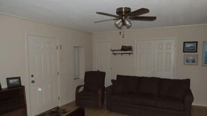Un salon avec un canapé marron foncé, une table basse en bois, un ventilateur de plafond et une porte dotée d’une petite fenêtre.