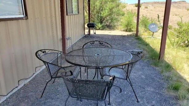Une terrasse extérieure couverte, avec une table ronde en métal et quatre chaises, une petite lampe de table et un panneau indicateur.