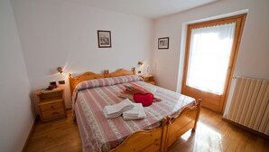 A bedroom with a wooden bed, two bedside tables, a window with curtains, and a radiator. - THREE-ROOM APARTAMENT  2 BATHROOM
BIKE TREKKING  MOUNTAIN VALFURVA STELVIO GAVIA (Santa Caterina)