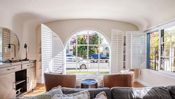 A cozy living room with a large arched window, leather sofas, a coffee table, and a wooden cabinet with a mirror. - Spanish Home: Steps to Beach & 2nd St
-Quiet part of Belmont Shores (Long Beach)