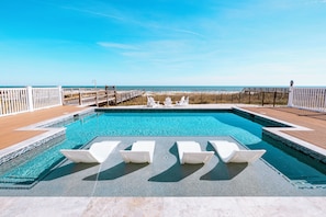 A pool with white lounge chairs, a wooden deck, and a view of the beach.