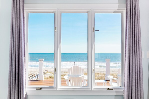 A room with a large window overlooking a beach, white chairs, and a wooden deck.