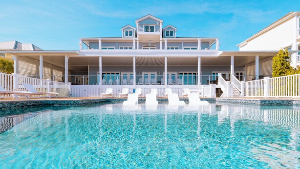 Une piscine bordée d’une terrasse blanche et dotée de chaises longues, un bâtiment à deux étages avec des balcons, et un ciel d’un bleu limpide.