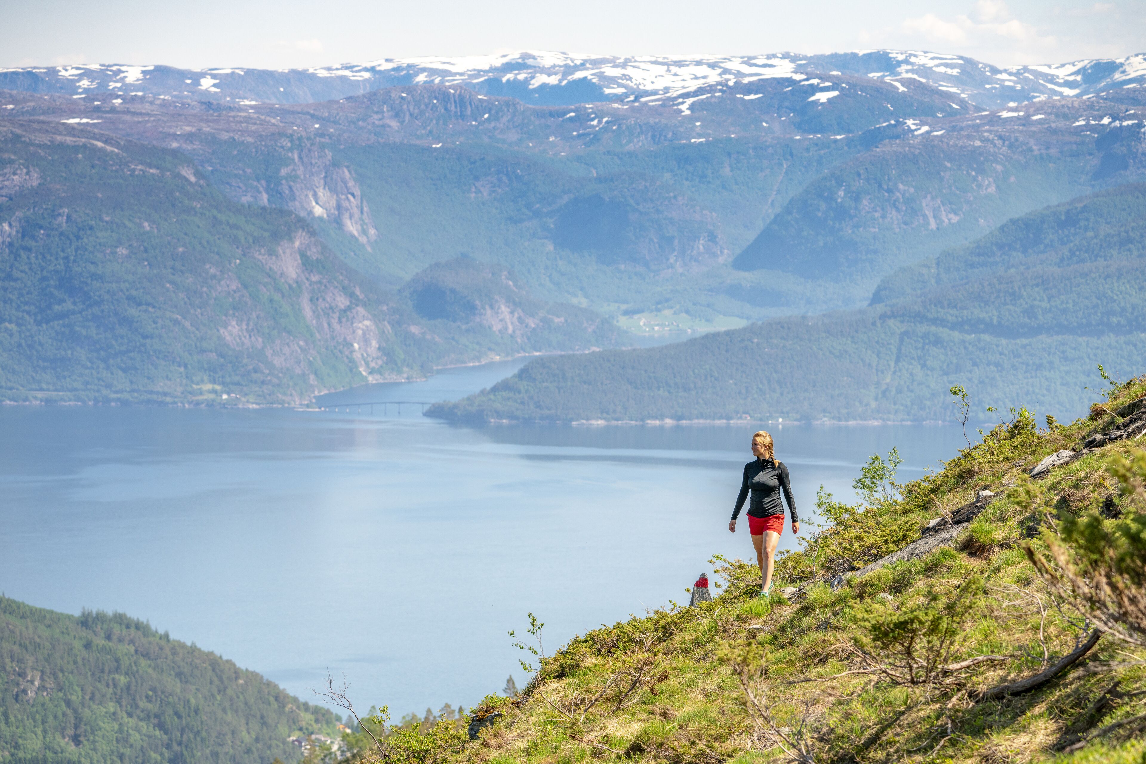 Una persona caminando por un sendero montañoso con un lago y montañas nevadas al fondo.