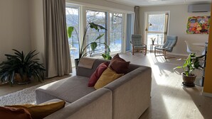 Una sala de estar con un sofá gris, una mesa de centro y un comedor con sillas. Hay ventanas amplias con cortinas, una planta en maceta y un cuadro en la pared.