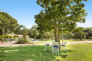 A grassy outdoor area with several white chairs and tables, surrounded by trees and a building in the background. - Ritz - East Hampton Cottage On The Water (East Hampton)