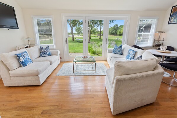 A living room with two white sofas, a glass coffee table, and a view of a garden through large sliding glass doors. - Ritz - East Hampton Cottage On The Water (East Hampton)