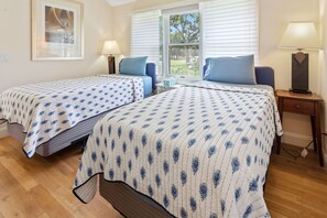 A room with two beds, each with a blue and white patterned bedspread, a window with blinds, a nightstand with a lamp, and a framed picture on the wall. - Ritz - East Hampton Cottage On The Water (East Hampton)