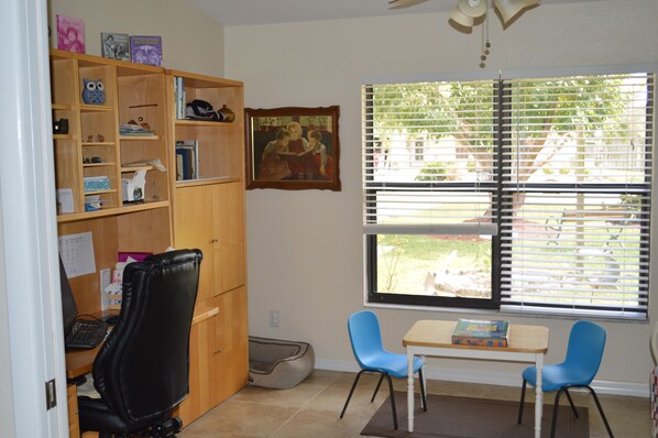 A room with a wooden desk, two blue chairs, a bookshelf, and a window with blinds.