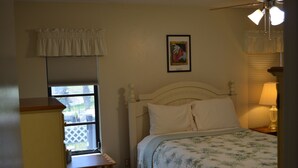 Une chambre à coucher avec un lit, une fenêtre avec des stores, un ventilateur de plafond et un tableau encadré au mur.