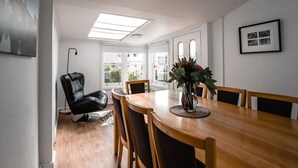 Een eethoek met een houten tafel, stoelen, een zwarte lederen fauteuil, een vaas met bloemen en een lijst met een foto aan de muur.