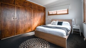 A bedroom with a large wooden wardrobe, a bed with white bedding, a bedside table, and a patterned ottoman.
