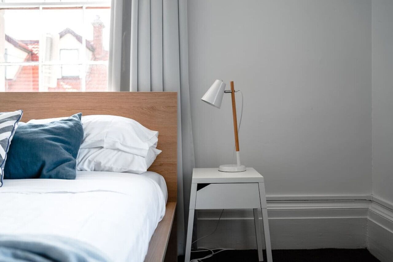 A neatly made bed with white and blue pillows, a white bedside table with a lamp, and a view of buildings through a window with curtains.