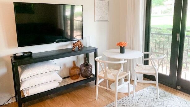 Ein modernes Wohnzimmer mit einem großen Flachbildfernseher an einer schwarzen Konsole, einem runden weißen Tisch mit zwei weißen Stühlen und einer Glastür, die zu einem Außenbereich führt.