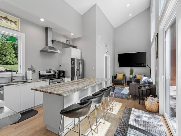 A modern kitchen with a central island, bar stools, a flat-screen TV, and a dining area with a sofa and coffee table. - Hot Tub+Fire Pit, On the River, WiFi, Grill, Cozy! (Burnsville)