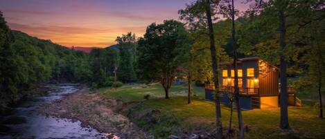 A cabin with a deck, surrounded by trees and a river.