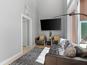 A modern living room with a sofa, a flat-screen TV, a patterned rug, and a door leading to another room. - Hot Tub+Fire Pit, On the River, WiFi, Grill, Cozy! (Burnsville)